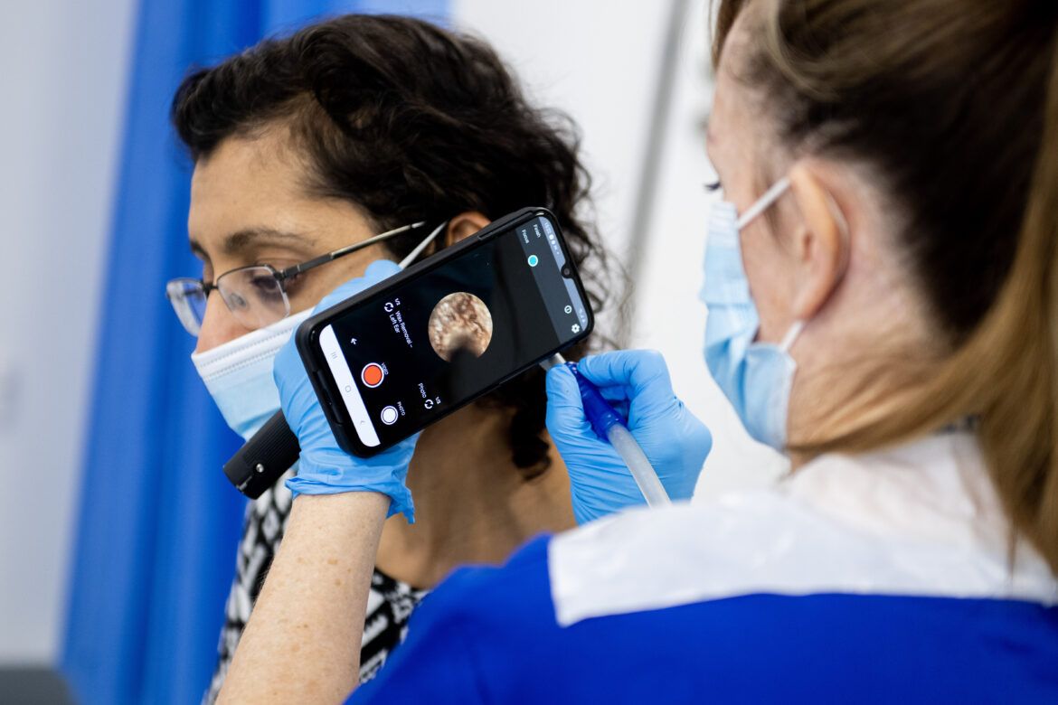 Two women in face coverings sitting in a medical setting. One is using a mobile phone with an app that can look into the patient's ear for assessment.
