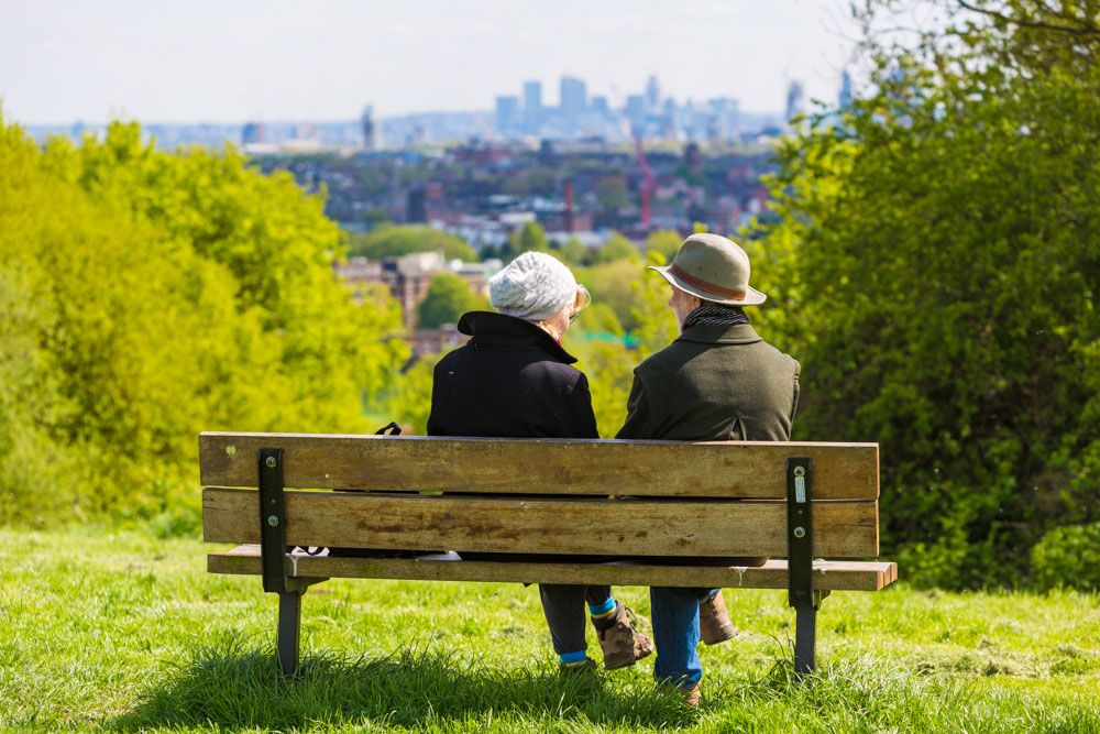 Two older people sitting on a bench in park on a sunny day. Photo is taken from the back so you are looking at the view with them.