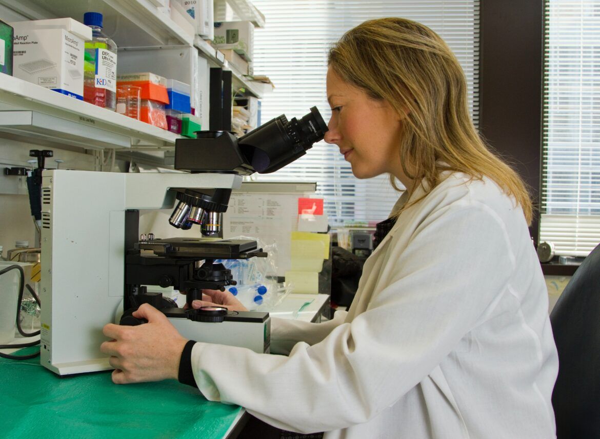 woman scientist with long blonde hair sitting at a research bench looking down a microscope. she is wearing a white lab coat.