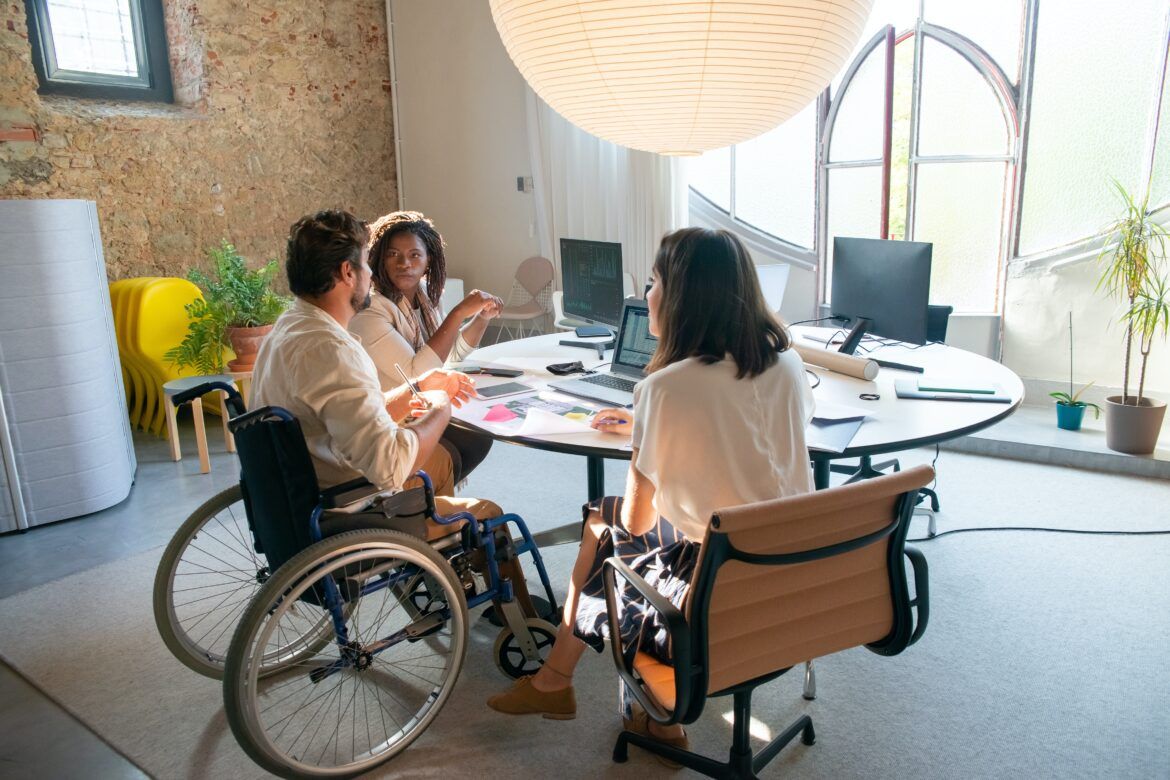 Photo of three people (one man and two women) sitting at a round table in an office. Man is in a wheelchair. 