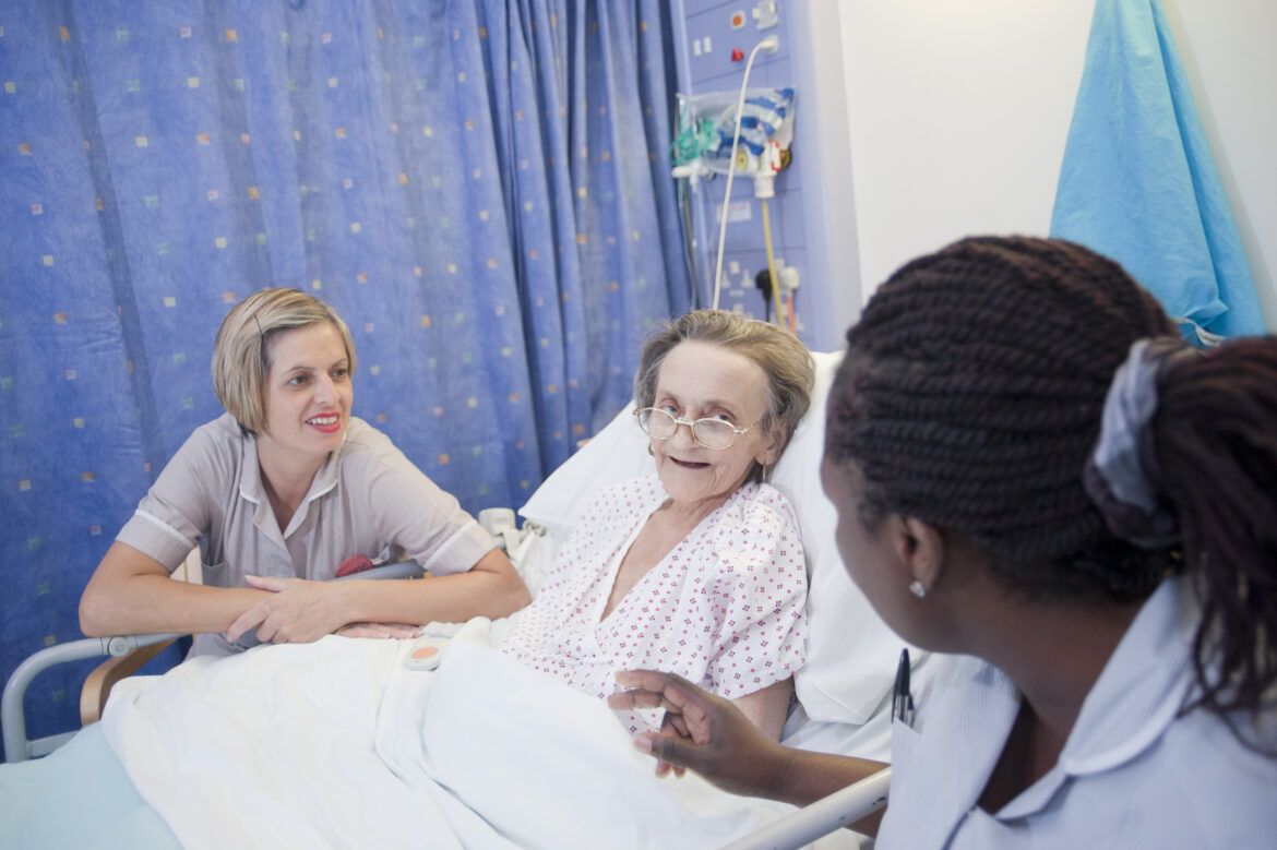 Photo of female elderly patient in hospital bed with two female healthcare professionals, all smiling and talking to eachother.